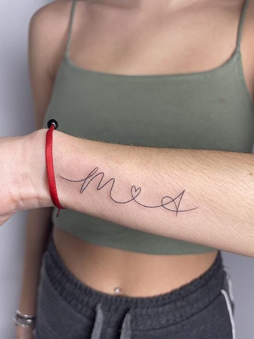 Tatuaje de iniciales de seres queridos entrelazados por un corazón en el antebrazo